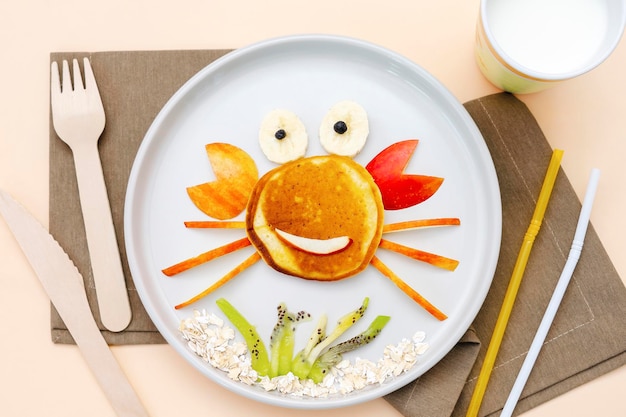 Funny crab face shape snack from pancakeapplesbananakiwi on plate Cute kids childrens baby's sweet dessert with milk healthy breakfastlunch food art on beige backgroundtop view flat lay