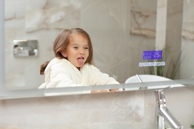 A funny crazy little girl in a bathrobe indulges in front of the mirror in bathroom and grimaces