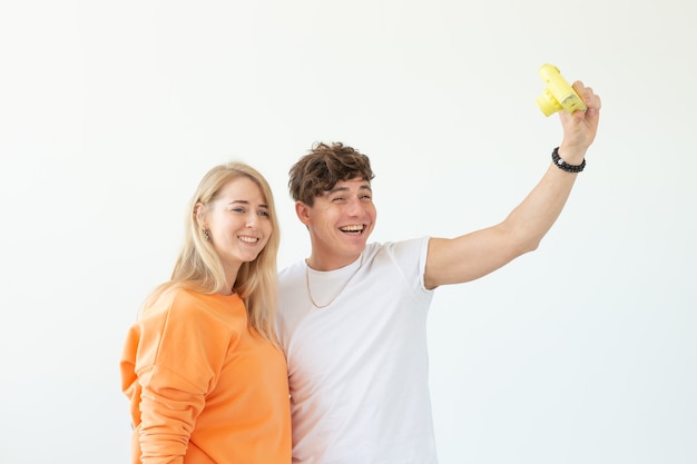 Funny crazy young couple blonde girl and a hipster guy taking a selfie on a vintage yellow film camera posing on a white wall. Concept of photography hobby. 