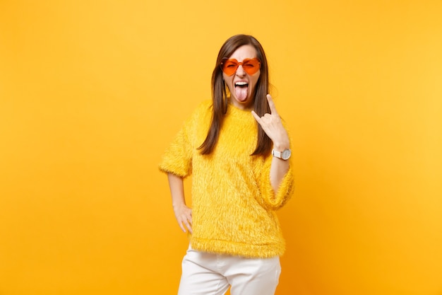 Photo funny crazy young woman in fur sweater and heart orange glasses screaming, showing rock-n-roll sign isolated on bright yellow background. people sincere emotions, lifestyle concept. advertising area.