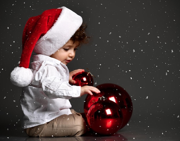 Funny curly baby in Santa hat playing with Christmas red balls on dark studio background New Year card first christmas snow