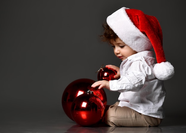 Funny curly kid in a Santa hat plays with New Year's red balls on a dark studio background New Year card first christmas