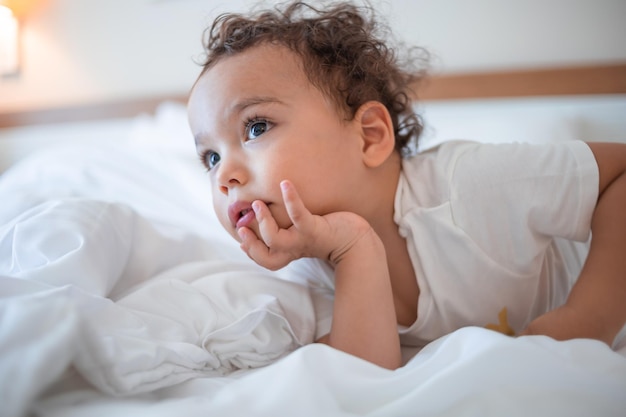 Funny curly toddler lying on the bed and attentively watching TV