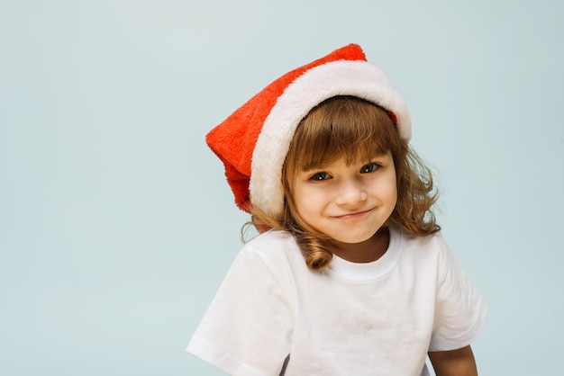 Funny cute girl in a santa hat stands on a blue background with copy space Concept of christmas and new year