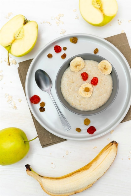 Funny cute kids childrens baby's healthy breakfast lunch oatmeal porridge in bowl look like bear face decorated with apple banana dried berry fruits dessert food art on white wooden table