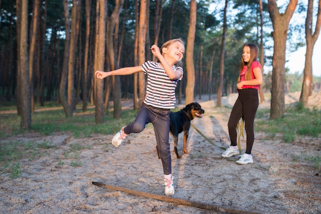 Funny cute little girl jumping over large old dry branch with wide smile on her face in dense pine forest on warm sunny summer bright morning, and sister with dog is standing in the background