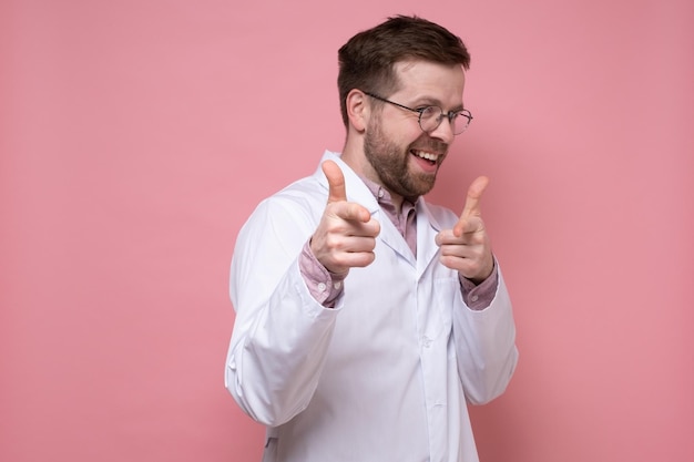 Funny doctor in a white coat is joking and smiling pointing at someone with fingers Pink background