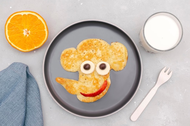 Funny elephant face shape snack from pancake on plate Cute kids childrens baby's sweet dessert healthy breakfastlunch food art with milkapplesorange on gray concrete backgroundtop view