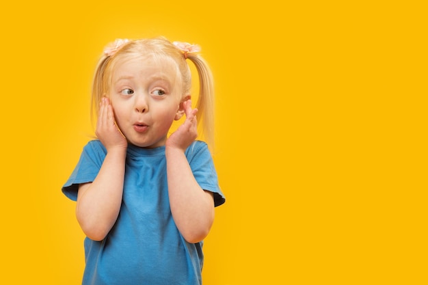 Funny fairhaired girl with two ponytails wears blue Tshirt and covers her ears looking away in surprise Yellow background Copy space