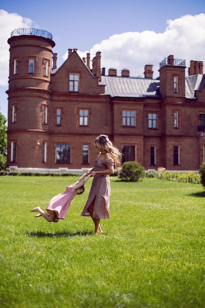 Funny family mom and daughter in pink dresses spinning through a green meadow against the background of the castle building in the summer heat