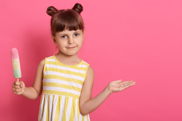 Funny female child spreading palms aside and looking directly at camera, posing isolated over pink background, having two knots, wearing striped summer dress.