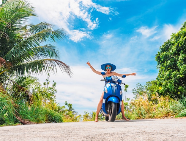 funny girl with opened hands riding a motorbike on a blue sky and green tropics background