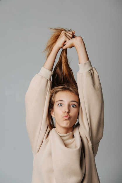 Photo funny grimacing teenage girl pulling her hair up, looking at the camera, making kissing lips. against grey background. in a warm beige longsleeve.