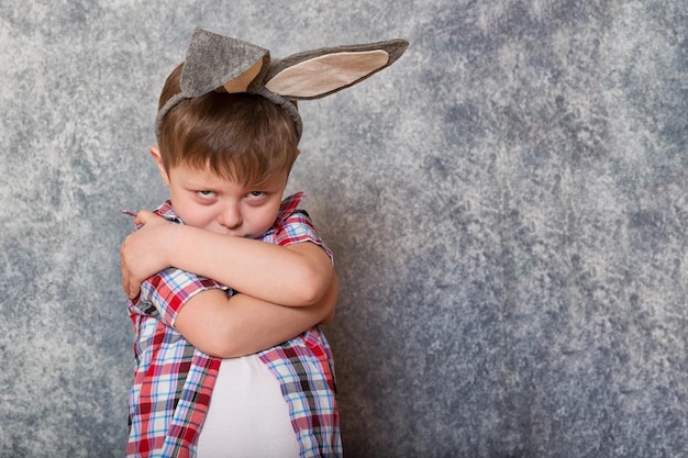 A funny grumpy little boy dressed in a red checkered shirt has a rabbit ears on his head