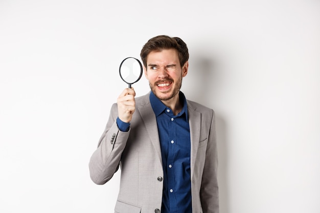 Funny guy in suit investigating or searching for something, looking through magnifying glass, standing on white background.