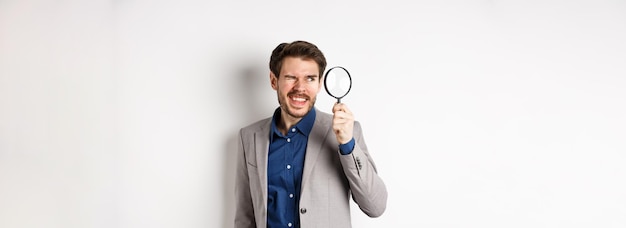 Funny guy in suit investigating or searching for something looking through magnifying glass standing