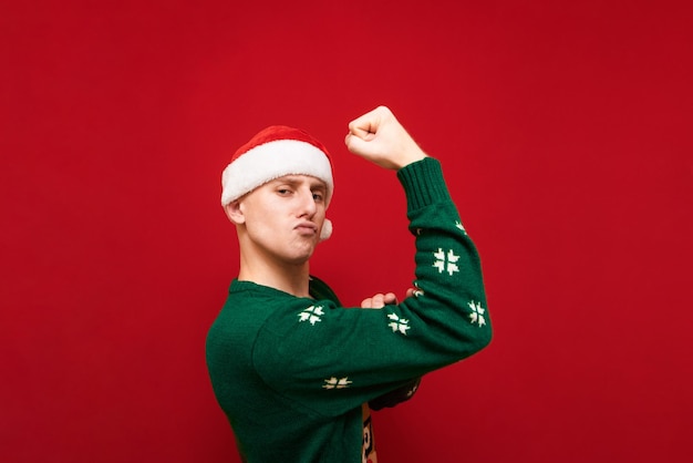 Funny guy with serious face stands on red background in christmas clothes and shows biceps