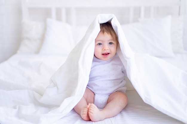 Funny happy baby girl looks out from under the blanket on the bed in white clothes and smiles a small child on a cotton bed at home woke up in the morning after sleeping