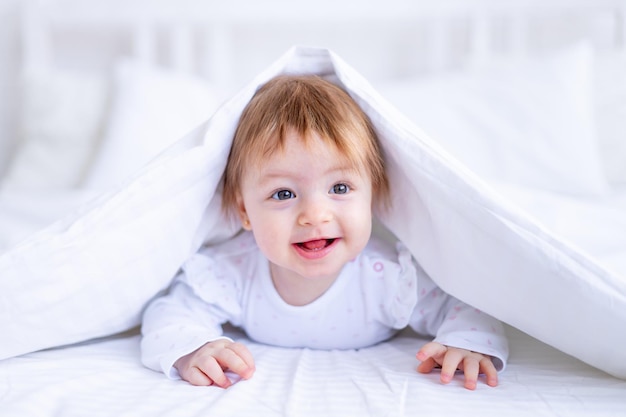 Funny happy baby girl looks out from under the blanket on the bed in white clothes and smiles a small child on a cotton bed at home woke up in the morning after sleeping