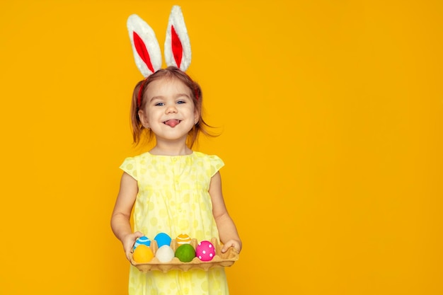 Funny happy baby girl with easter eggs and bunny ears on yellow background