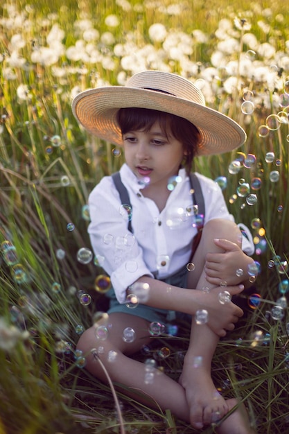 Funny happy a beautiful boy child sit in hat on a field with white dandelions at sunset in summer soap bubbles are flying