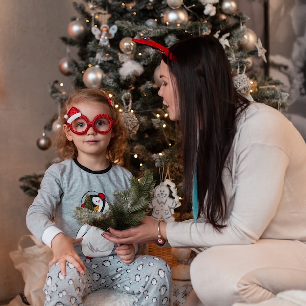 Funny happy family, mom and little daughter child in fashionable pajamas sit near Christmas trees with lights. Winter holidays at home