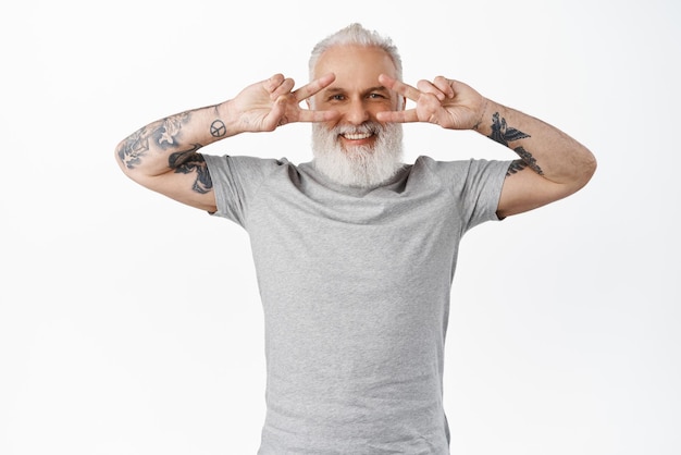 Funny and happy senior guy with tattoos showing disco peace vsigns near eyes smiling and looking positive at camera standing in gray tshirt against white background