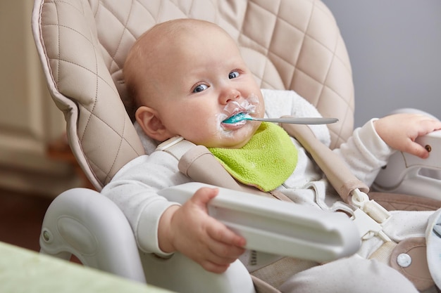 Funny kid is sitting with a spoon in his mouth