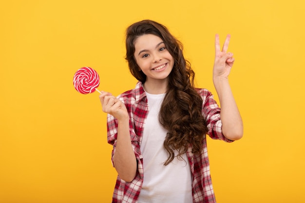 Funny kid with long curly hair hold lollipop caramel candy on yellow background show peace gesture, childhood.