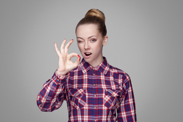 Funny kissing beautiful girl with pink checkered shirt, collected bun hairstyle and makeup standing and showing Ok sign and looking at camera with wink. indoor shot, isolated on gray background.