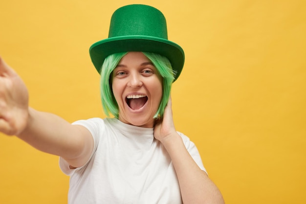 Funny laughing woman with green hair wearing leprechaun hat posing isolated over yellow background making point of view photo exclaiming happily creating content for blog