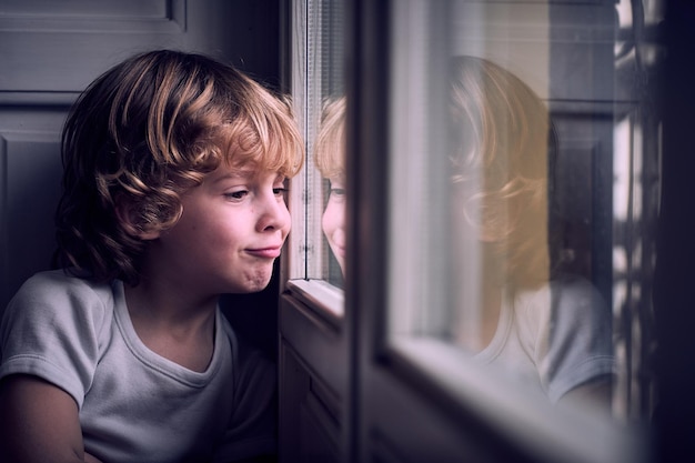 Funny little bot with curly blond hair in white t shirt pouting lips and looking our window with interest at home