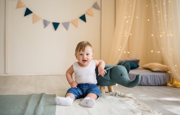 Funny little boy in a white bodysuit and jeans is sitting on the floor in the children's room with toys and looking at the camera