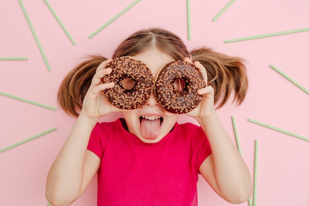 Funny little cheerful girl on a pink background with chocolate donuts green cocktail tubes Top view