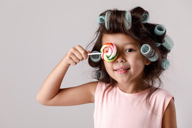 Funny little child girl in pink dress and hair curlers holding lollipop on gray background.