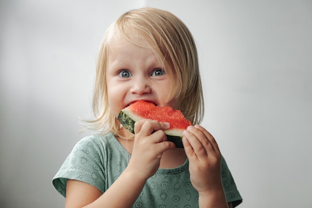 Funny little girl eating watermelon close-up. Cute child with watermelon indoors. Concept of healthy food, summertime. Copy space