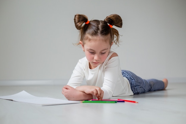 Funny little girl is lying on the wooden floor and drawing in an album with colored pencils