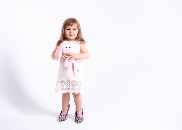 Photo funny little girl in white dress trying her mother shoes with heels on white surface.