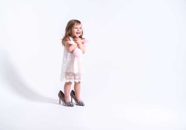 Photo funny little girl in white dress trying her mother shoes with heels on white wall