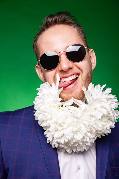 Funny male in suit and sunglasses licking petal of flowers while having fun in studio on green background and looking at camera