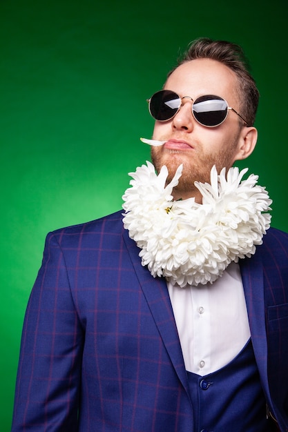 Funny male in suit and sunglasses licking petal of flowers while having fun in studio on green background and looking at camera