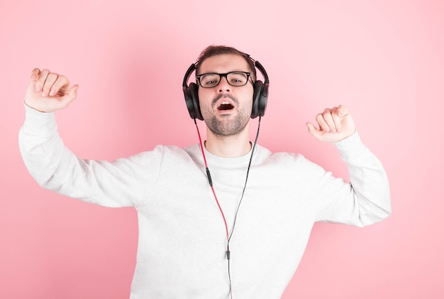 A funny man with a beard and a white sweatshirt listens to music with headphones, sings and dances on a pink background.