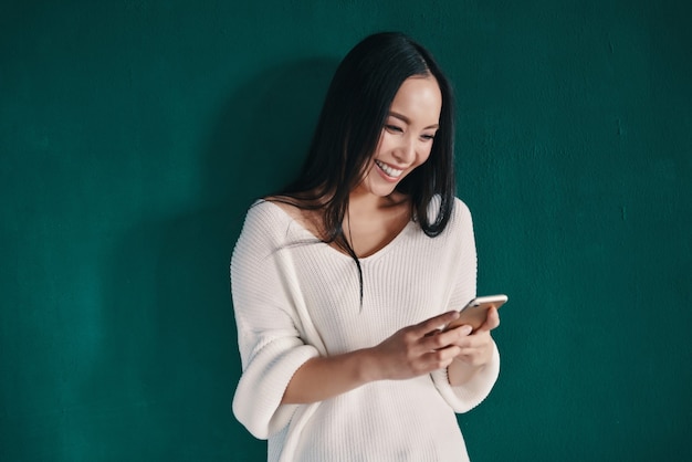 Funny message. Beautiful young woman using smart phone and smiling while standing against green wall