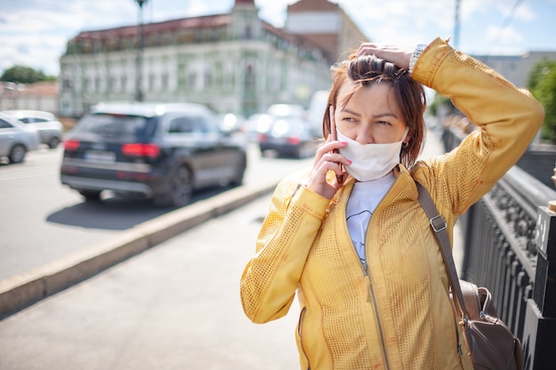 Funny middle-aged woman in a white protective mask talking on a smartphone while walking around the city on a warm spring day