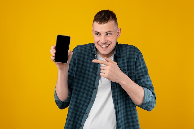 Funny millennial man showing cellphone blank screen on yellow background