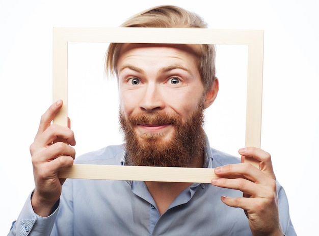 Funny picture. Young man wearing beard holding picture frame in front of his face and fun hamming, isolated on white.