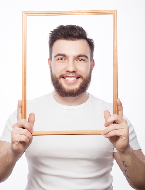 Funny picture. Young man wearing beard holding picture frame in front of his face and fun hamming, isolated on white.