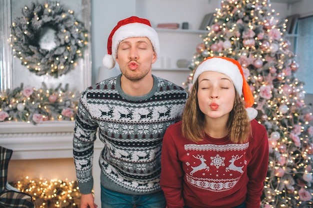 Funny picture of young man and woman stands and poses. They keeps eyes closed. People's lips are in kiss shape. There are Christmas tree and fireplace behind them.