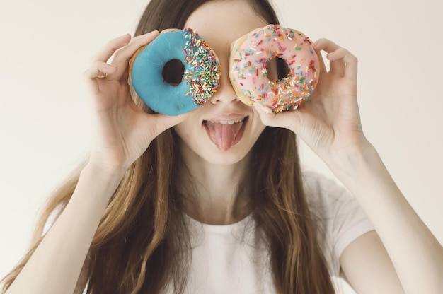 Funny portrait of a young female model holding blue and pink donuts near eyes and showing a tongue