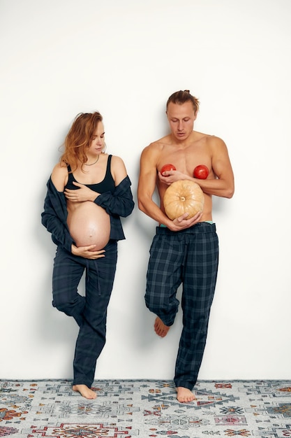 Funny pregnant girl with her husband play Pregnant woman holds her belly and her husband holds a pumpkin instead of his belly and tomatoes at his chest